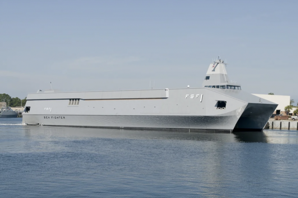 A sleek, gray naval ship, marked "FSF-1," sails on a calm blue sea near a port. The vessel has a modern design with a substantial deck and streamlined structure. Clear sky and distant land are visible in the background.