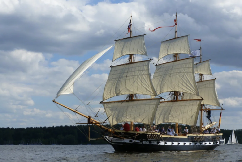 A large sailboat with multiple masts and cream-colored sails is navigating on a body of water under a cloudy sky. The vessel is filled with people, and the shoreline with trees is visible in the background.