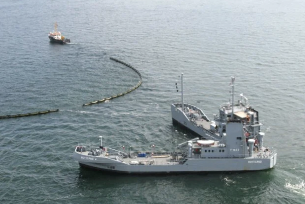 Aerial view of two ships on a body of water. The larger ship, labeled "ROTTERDAM," is in the foreground, while a smaller vessel follows behind. A floating barrier or boom extends in the water between them.