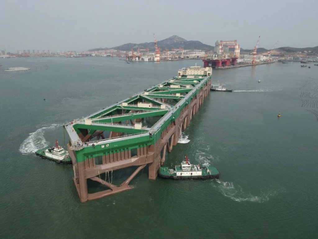 Aerial view of a large green and brown structure being transported on water by several tugboats. In the background, there's a shipyard with cranes and a mountain visible. The water is calm under a clear sky.