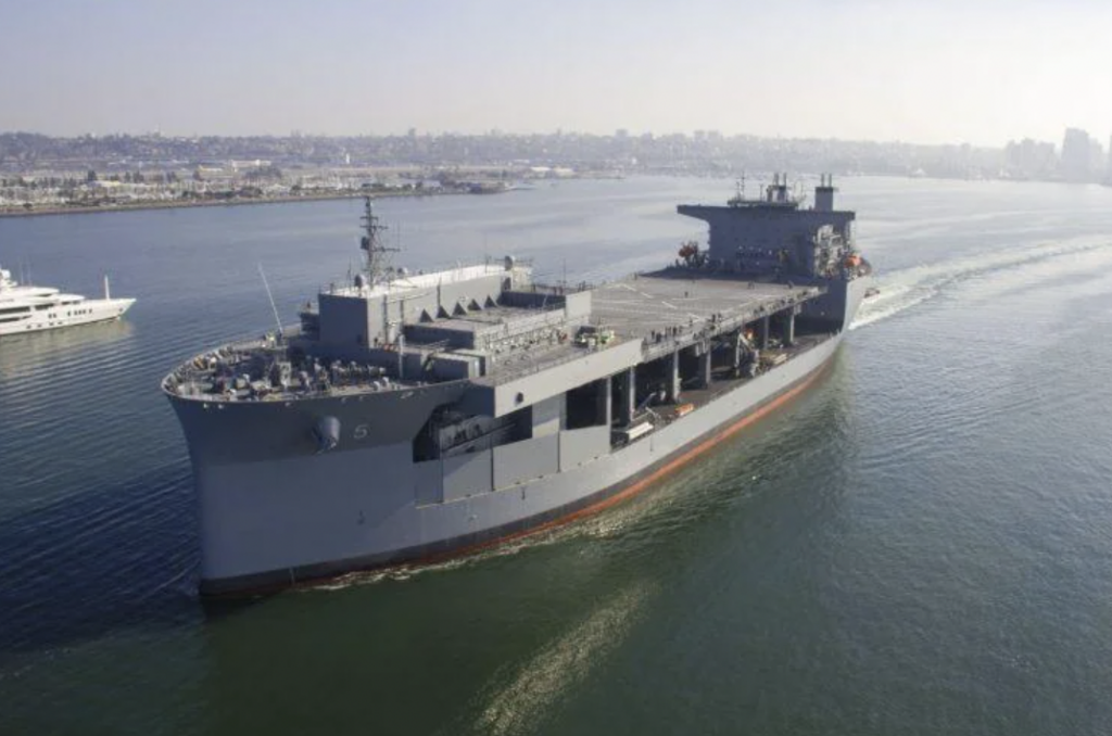 A large gray military ship sails through calm waters, with a small group of people visible on deck. The ship is near a coast, where a cityscape with modern buildings is visible in the background. A white yacht is seen to the left.