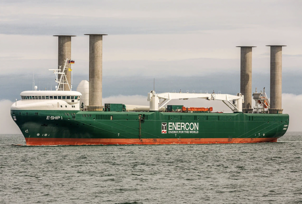 Green and white cargo ship labeled "ENERCON" with distinctive tall, cylindrical structures on deck, sailing on a calm sea. The ship's hull reads "E-SHIP 1." The sky is overcast.