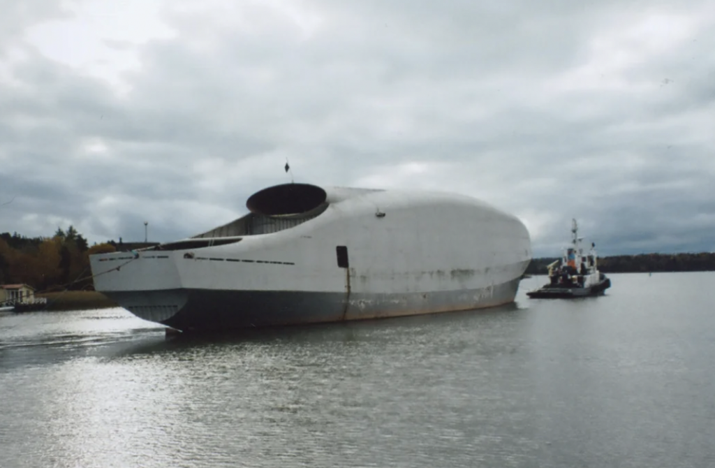 A large, uniquely shaped cargo ship with a rounded, covered front is being guided by a small tugboat on a calm body of water. The sky is overcast, and trees are visible in the background.