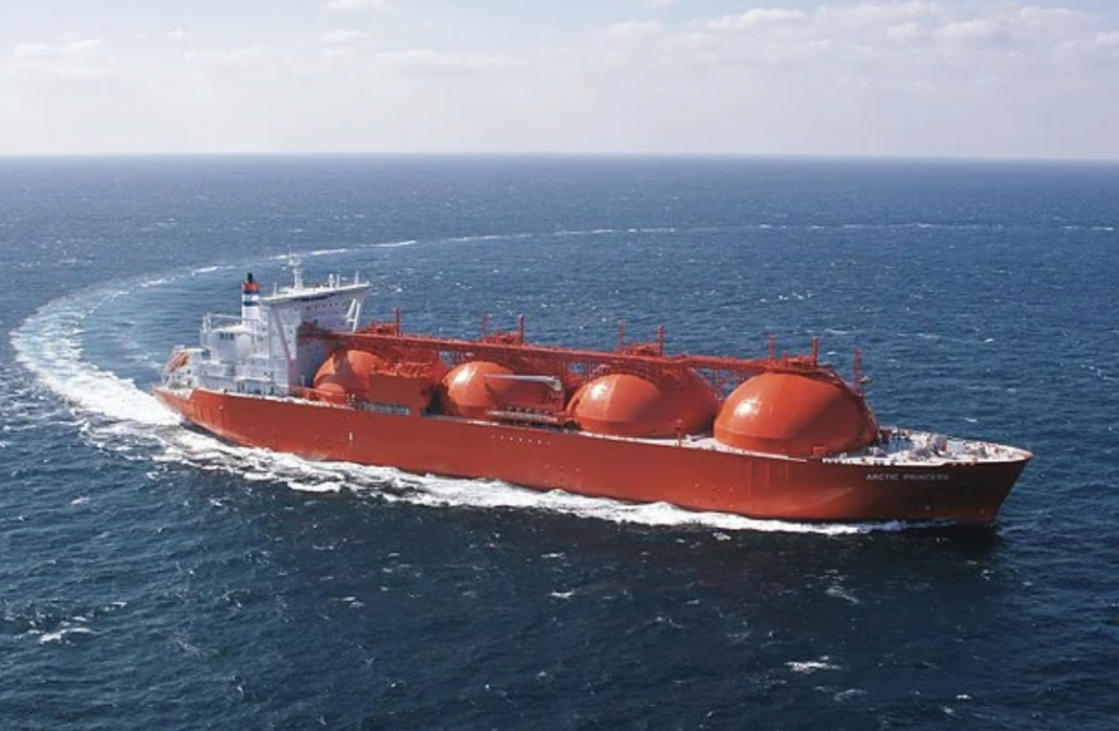 A large red LNG tanker with spherical storage tanks sails through deep blue ocean waters under a lightly clouded sky. The ship creates a white wake as it moves forward.