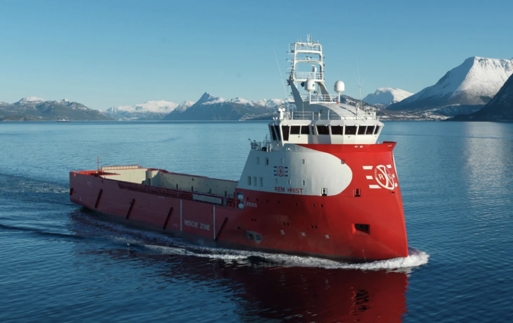 A large, red cargo ship with white accents sails on a calm body of water, surrounded by snow-capped mountains under a clear blue sky. The ship's name and logo are visible on its side.