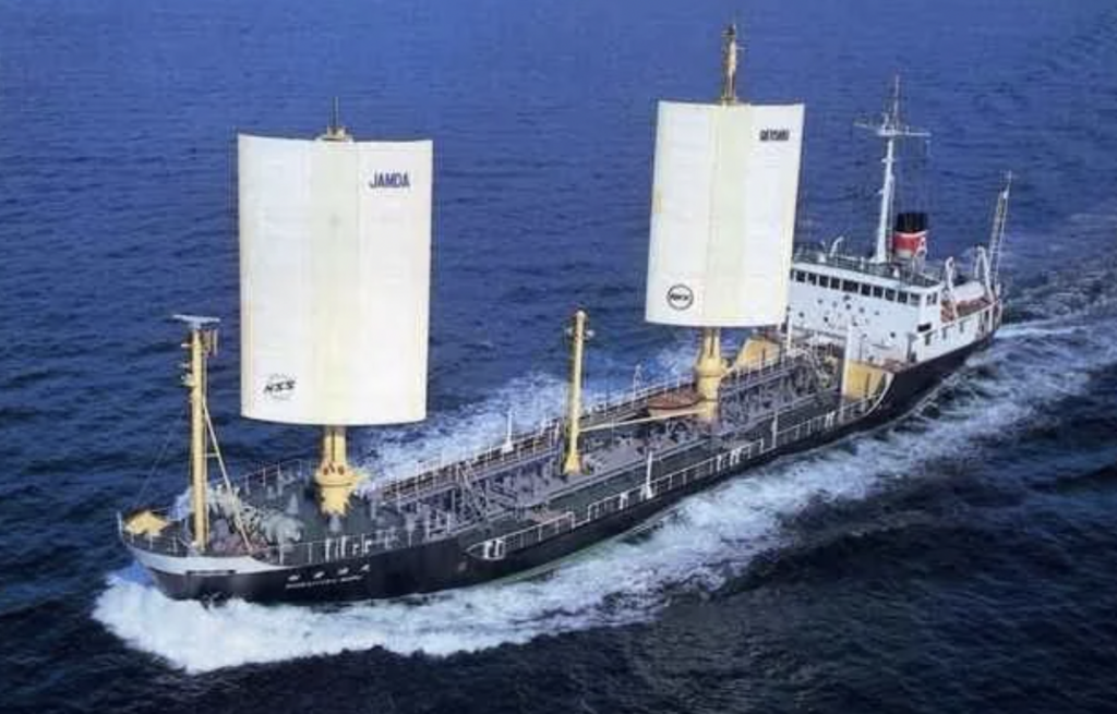 A cargo ship equipped with two large, vertical sails navigates through open blue waters. The vessel is surrounded by waves, showcasing its movement. The ship bears markings with the words "JAMDA" and "OMIYAM," visible on the sails.