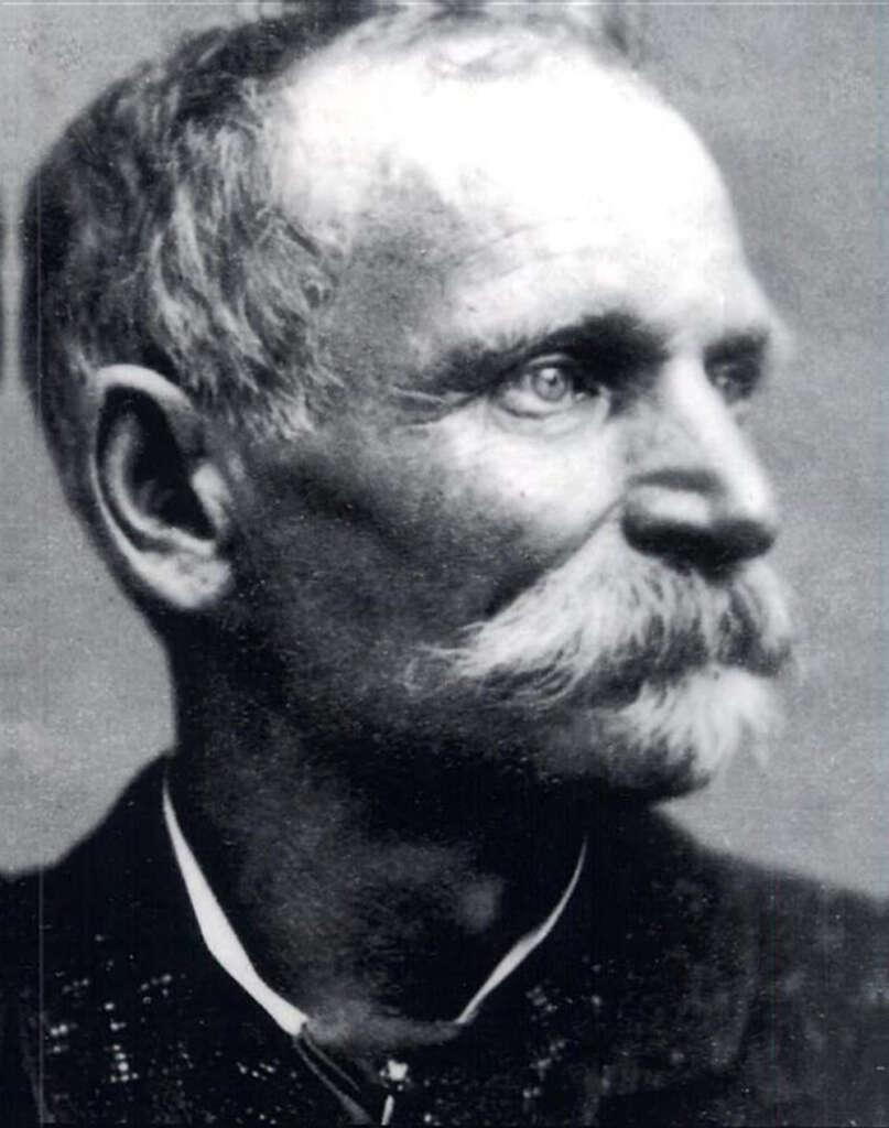 Black and white portrait of an older man with a prominent mustache, looking slightly to the right. The man has short hair and is wearing a dark suit with a visible collared shirt underneath. The background is plain and blurred.