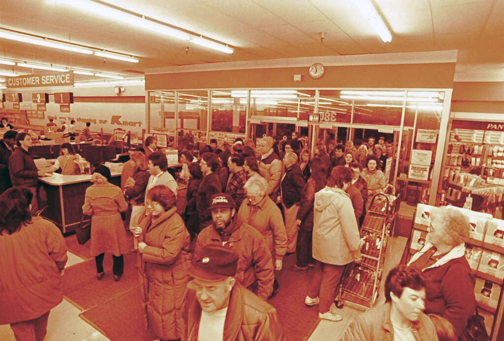 A large group of people crowd inside a store entrance, waiting near the customer service area. The scene appears to be bustling, with many shoppers wearing coats and some standing in line. Fluorescent lights illuminate the store interior.