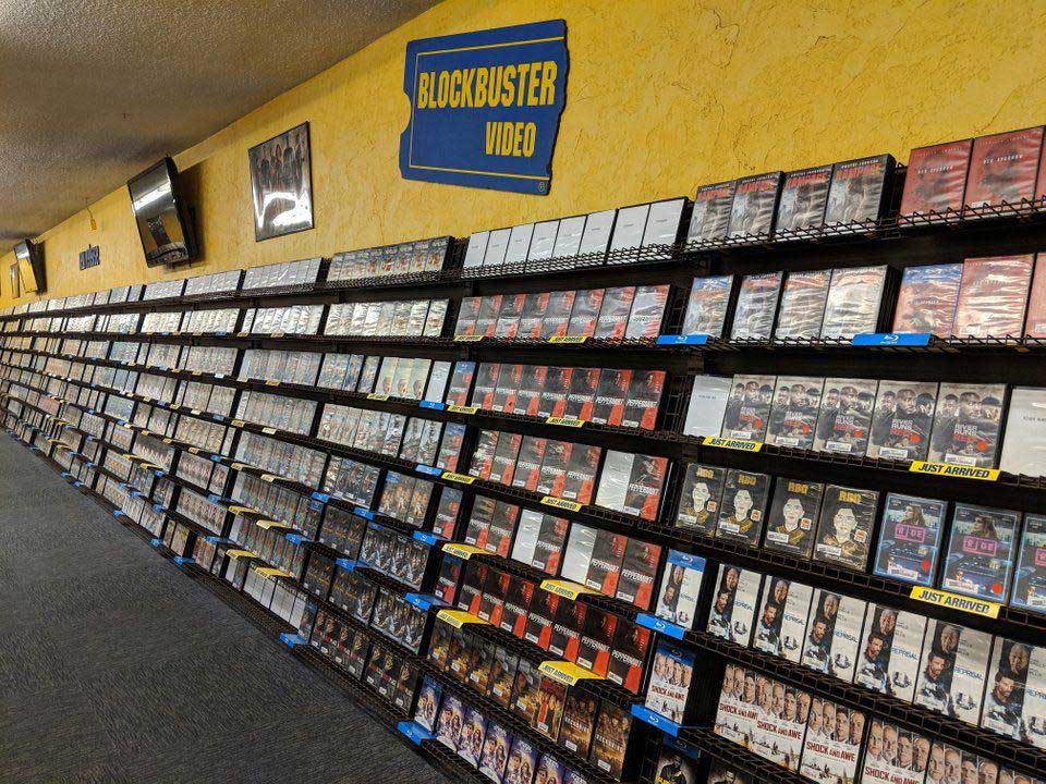 A long aisle in a Blockbuster Video store with shelves filled with DVDs and video cases. A yellow wall and blue Blockbuster Video sign are visible above the shelves. The carpeted floor runs parallel to the shelves.