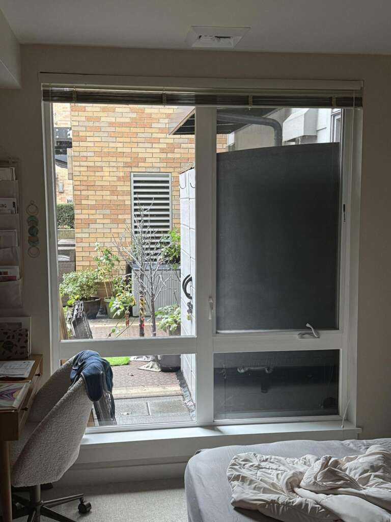 View of a window overlooking a small outdoor patio with potted plants. A desk with a chair is on the left, while a bed with a rumpled blanket is on the right. The patio is enclosed by a brick wall.