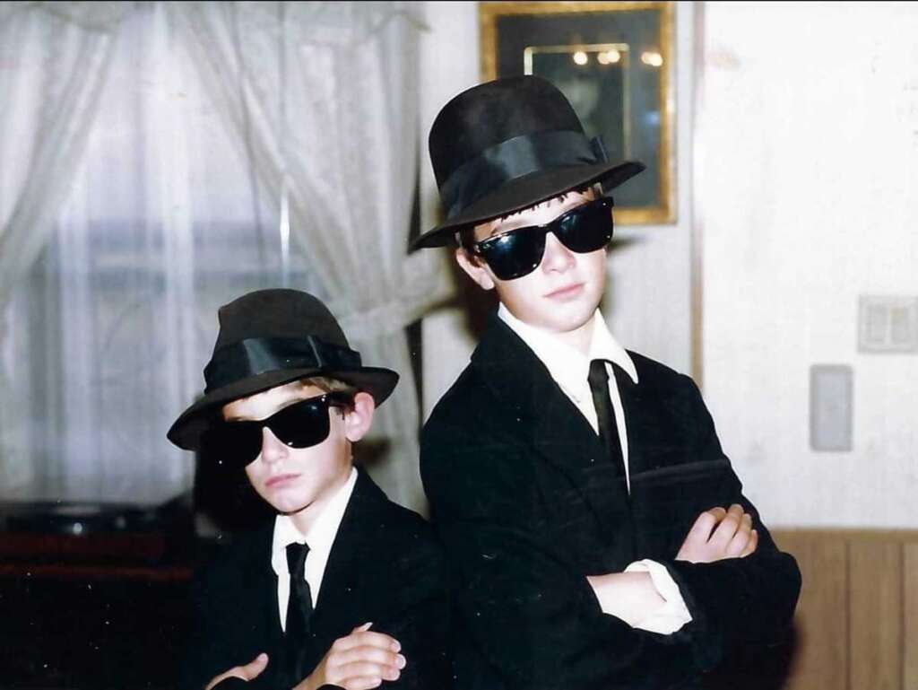 Two children dressed in black suits, hats, and sunglasses stand confidently with arms folded. They're inside a room with a window and curtains in the background, resembling characters from a classic film.