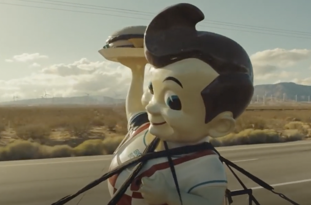 A large mascot statue of a smiling boy holding a hamburger is strapped onto a vehicle. The desert landscape with a few shrubs and wind turbines can be seen in the background under a cloudy sky.