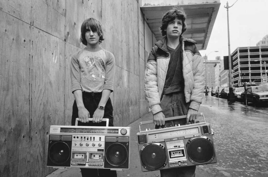 Two young individuals stand on a city street holding large boomboxes. They wear casual clothing, and the background features a wooden wall and parked cars. The scene has a vintage, urban feel.