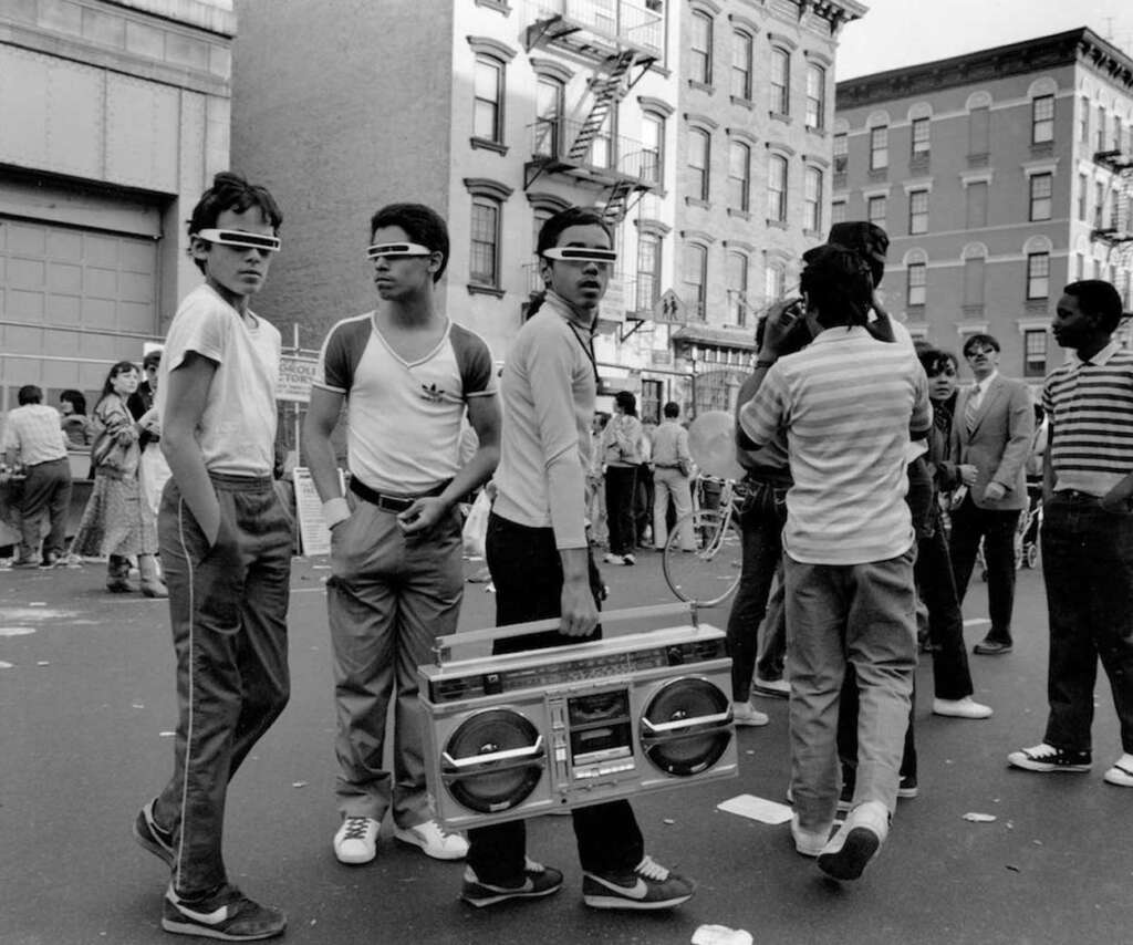 A group of young men in casual 1980s attire stands on an urban street wearing futuristic sunglasses. One holds a large boombox. Other people are gathered in the background near apartment buildings.