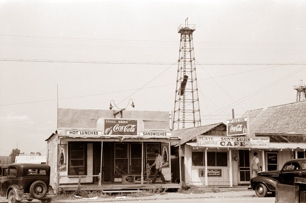 A vintage street scene featuring two small shops: one advertising Coca-Cola and sandwiches, and the other a café. A tall oil derrick rises in the background. Classic cars are parked in front, with a couple of people nearby.