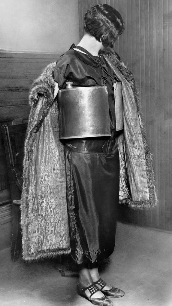 1920s woman in a dress and fur coat concealing metal containers under her arms, likely as a form of smuggling during Prohibition. The image is in black and white, capturing the fashion and clandestine activity of the era.