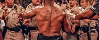 A shirtless man with a muscular back faces several uniformed officers who appear to be stopping him. The scene is tense, with officers extending their arms toward him. The setting is a crowded area, possibly an event or arena.