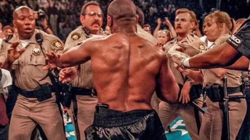 A shirtless man with a muscular back faces several uniformed officers who appear to be stopping him. The scene is tense, with officers extending their arms toward him. The setting is a crowded area, possibly an event or arena.