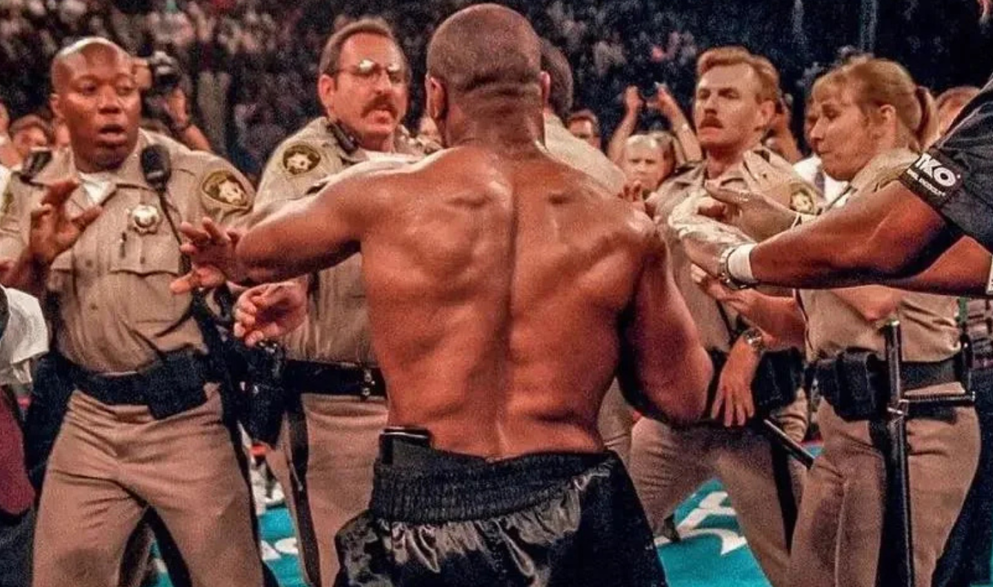 A shirtless man with a muscular back faces several uniformed officers who appear to be stopping him. The scene is tense, with officers extending their arms toward him. The setting is a crowded area, possibly an event or arena.