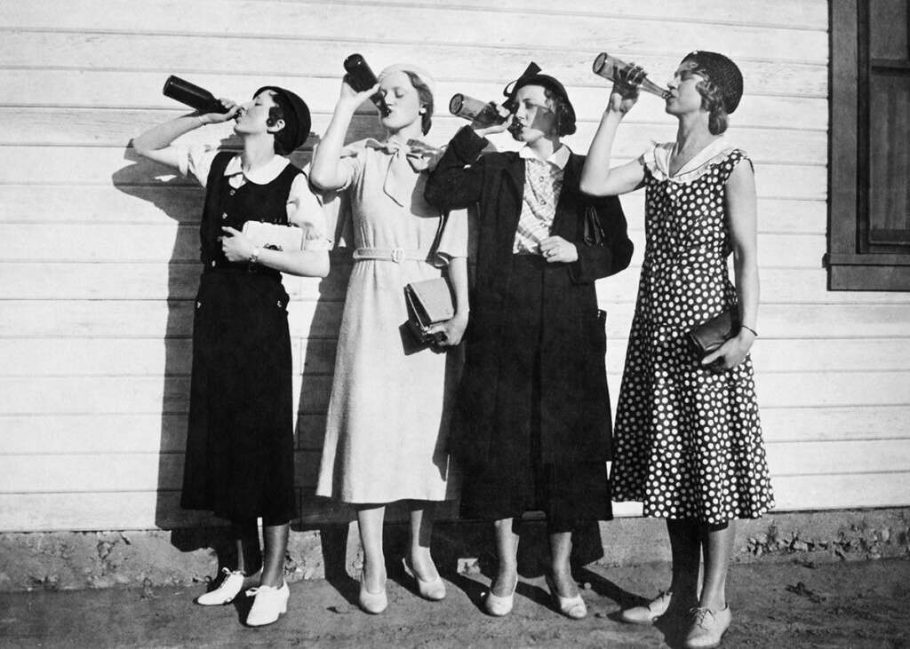 Four women in vintage clothing stand against a wooden wall, each drinking from dark bottles. They wear hats and dresses, with one in polka dots, enjoying a sunny day. The scene has a nostalgic, early 20th-century feel.