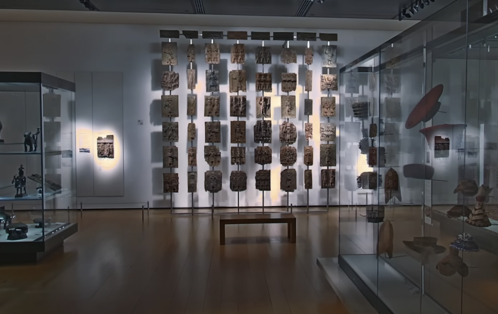 Museum exhibit featuring a large display of ancient artifacts positioned on a wall, with glass cases containing various sculptures and objects nearby. The room has wooden floors and is softly lit, highlighting the historical pieces.