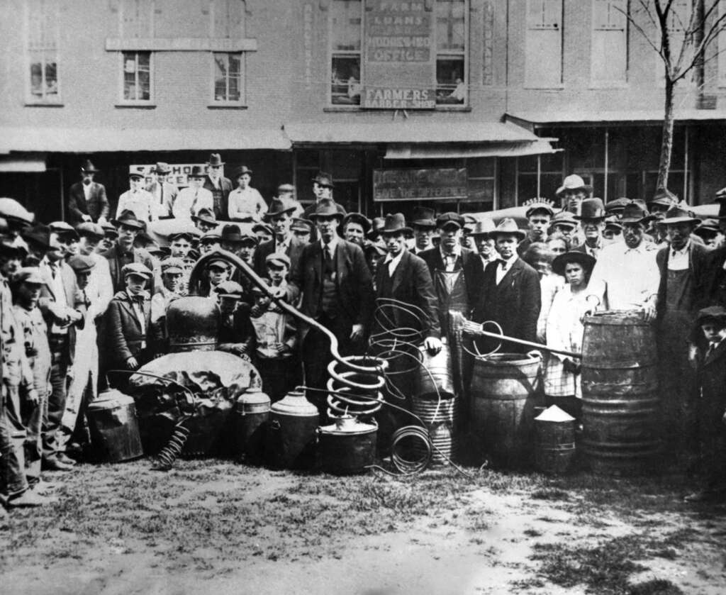 A historical black and white photo showing a large group of adults and children gathered in front of a building. They are surrounded by barrels, coiled tubing, and containers, possibly related to distillation or brewing equipment.