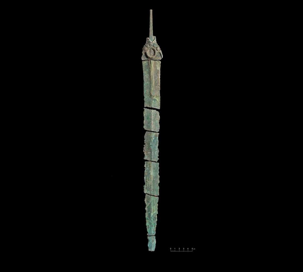 Ancient, oxidized bronze artifact resembling a long, narrow blade with a pointed tip, displayed vertically against a black background. The surface is textured and segmented, with visible wear and green patina. A measurement scale is at the bottom.