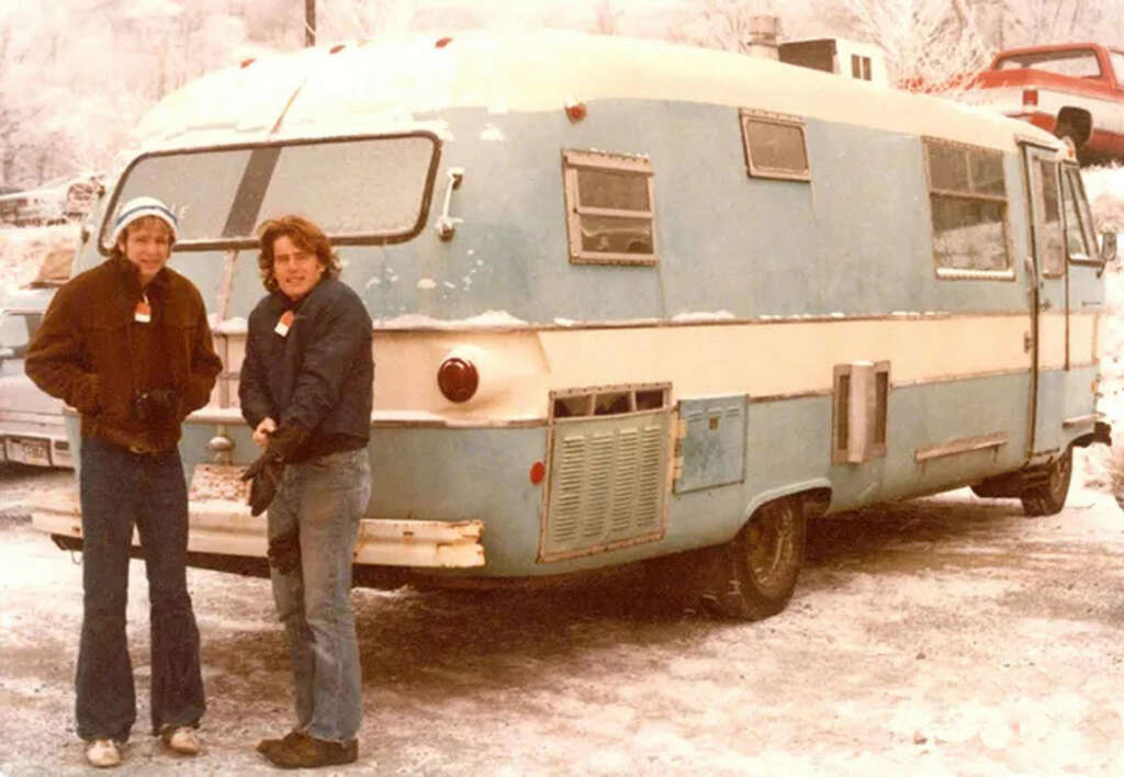 Two people stand in front of a vintage blue and white camper van on a snowy road. They're wearing winter jackets and hats, indicating cold weather. The camper has snow on its roof and windshield. Cars and snowy trees are in the background.