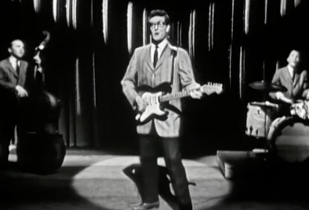 A black-and-white image of a band performing on stage. The central figure is playing an electric guitar, flanked by a double bassist on the left and a drummer on the right, both also playing. The backdrop features vertical striped drapes.