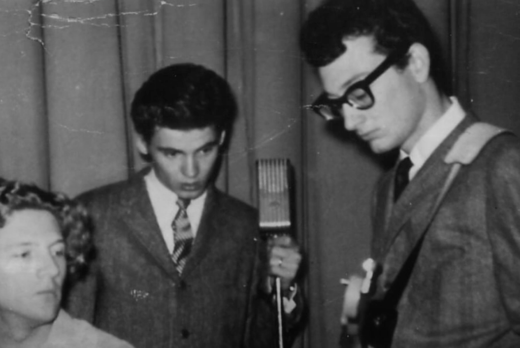 A black and white photo shows three men in suits gathered around a vintage microphone. One is seated with curly hair, another stands close to the microphone, and the third, wearing glasses, holds a guitar. The backdrop is a plain curtain.