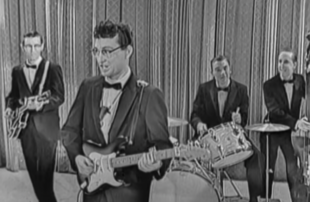 A black and white image shows a band of four men in suits playing musical instruments. The guitarist in front is singing, with other members playing electric guitar, drums, and bass guitar in the background, standing before a curtain.