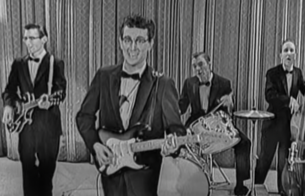 A black-and-white photo of a band performing. The lead guitarist, wearing glasses and a tuxedo, smiles at the camera. Behind him, a drummer, a bassist, and another musician, all in tuxedos, are playing their instruments. They are on a stage with curtains.