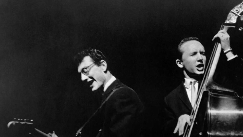 Two musicians performing on stage in black and white. The one on the left plays an electric guitar, wearing glasses and a suit. The musician on the right plays an upright bass, dressed in a suit and tie, both appearing energetic and focused.