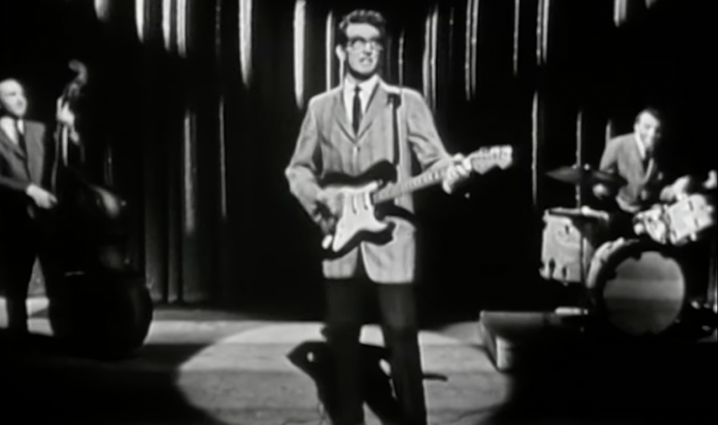 A black-and-white photo shows a musician playing an electric guitar on stage. He's flanked by a double bassist on the left and a drummer on the right, with a curtain backdrop and spotlight highlighting the performers.