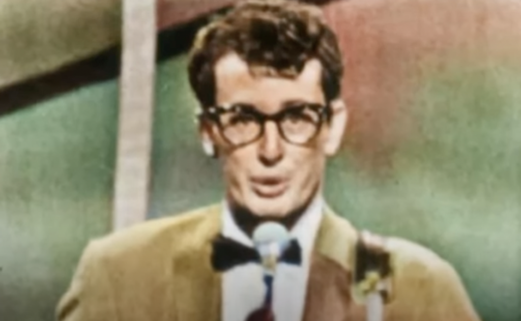 A man with black-rimmed glasses and a bow tie sings into a microphone. He wears a brown suit jacket. The background is colorful and blurred.