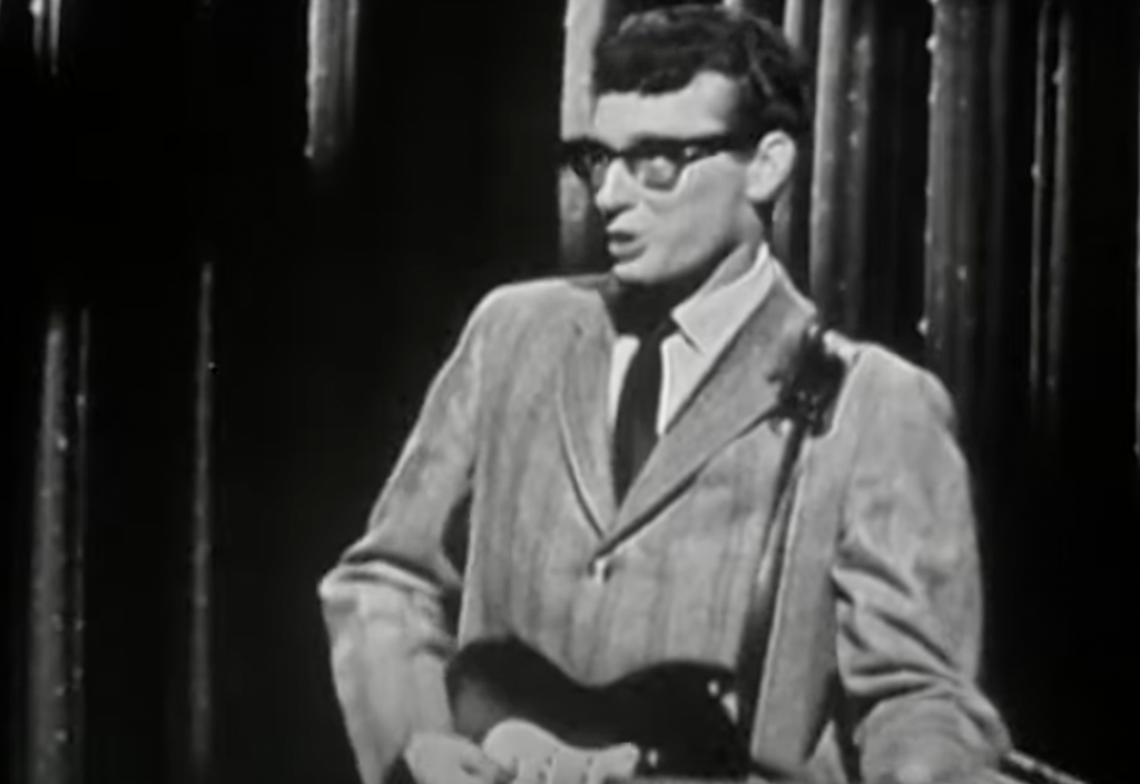 A man in glasses plays an electric guitar on stage. He is wearing a suit and tie, and the background is dark with thin vertical lights. This image is in black and white, suggesting a vintage or classic performance.