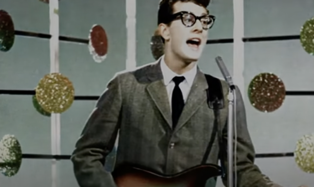 A man in a suit and glasses sings into a microphone while playing an electric guitar. The background features a pattern of large, colorful circles.