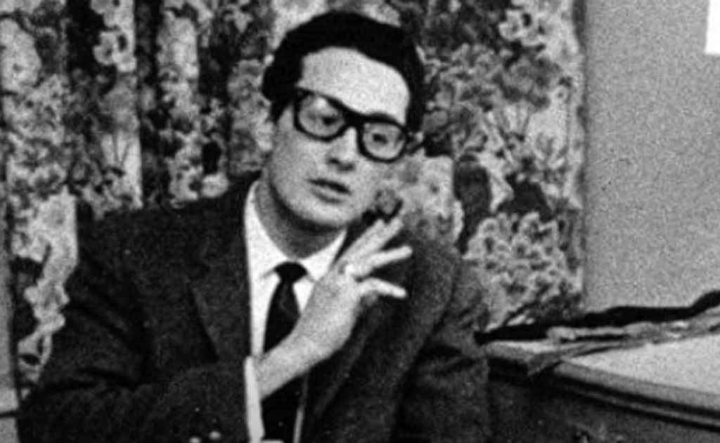 A black-and-white photo of a man wearing glasses and a suit, seated indoors. He gestures with one hand raised near his face. The background features a floral curtain and a portion of a table.