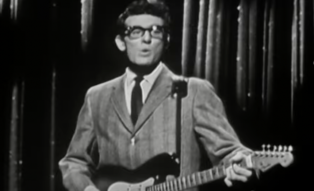 A man in a suit and glasses plays an electric guitar on stage. The image is in black and white and has a vintage appearance, with curtains visible in the background.