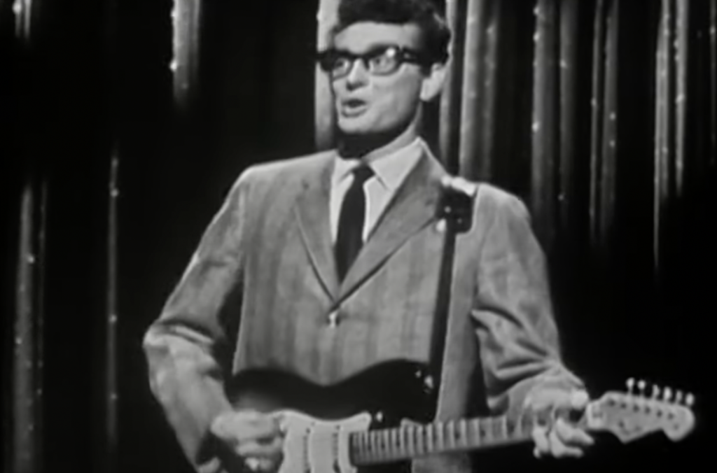 Black and white photo of a man in a suit and glasses playing an electric guitar in front of a curtain.