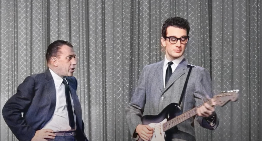 A man in a suit and dark glasses plays an electric guitar while another man in a suit stands to the side, looking at him. They are on a stage with a patterned curtain backdrop.