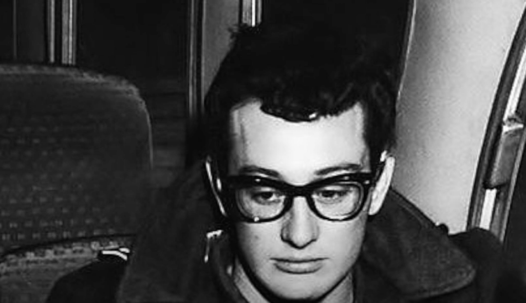 A person with dark hair, wearing thick-framed glasses and a jacket, sits in a bus or train seat. The mood appears contemplative. The background shows part of another seat and a window. The image is in black and white.