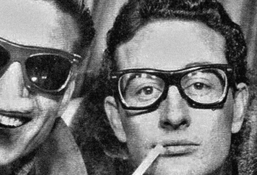 Two people wearing glasses, one with dark frames and a cigarette in their mouth, are smiling. The image has a vintage, black and white appearance.