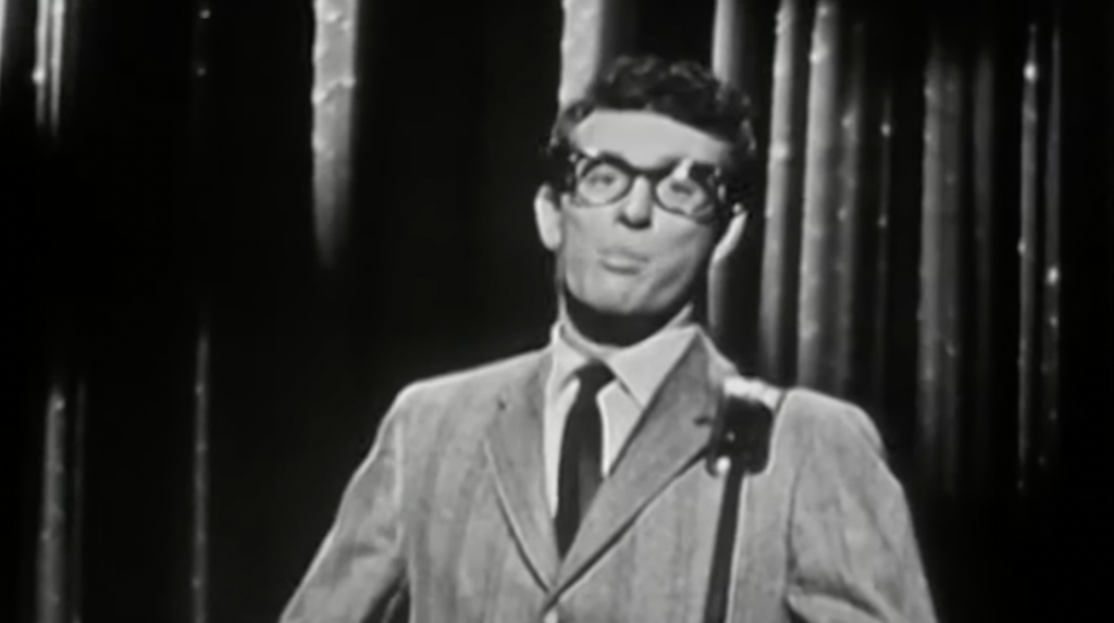 A man in a suit and glasses sings into a microphone on a dimly lit stage, with vertical lights in the background. The image is in black and white, evoking a vintage or retro atmosphere.