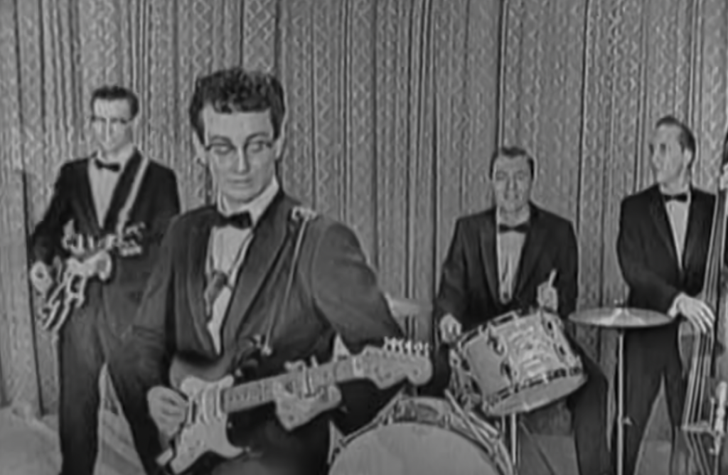 A black and white photo of a four-member band performing. The lead guitarist stands at the front, wearing glasses, and the drummer is in the background. All members are dressed in suits and bow ties, playing electric guitars and drums.