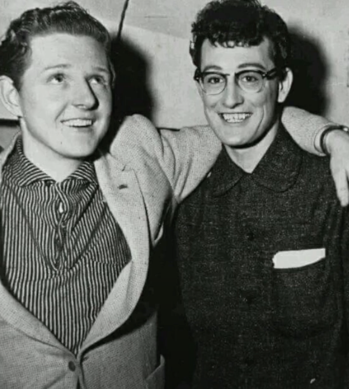 Two men in a black and white photo, both smiling. The man on the left wears a light suit and striped shirt, with one hand in his pocket. The man on the right wears glasses and a dark jacket with a pocket square. His arm is around the other man's shoulders.