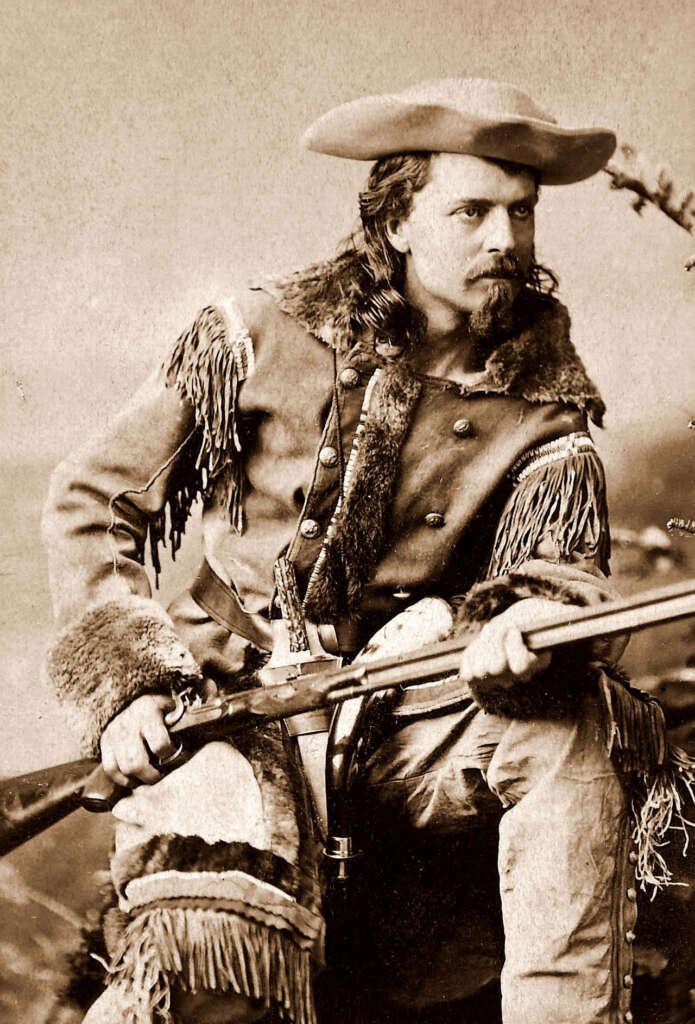 A person in 19th-century frontier attire with a wide-brimmed hat and tassel-lined jacket holds a long rifle. The sepia-toned image gives a historical feel. The individual sits with a serious expression, embodying a Wild West explorer.