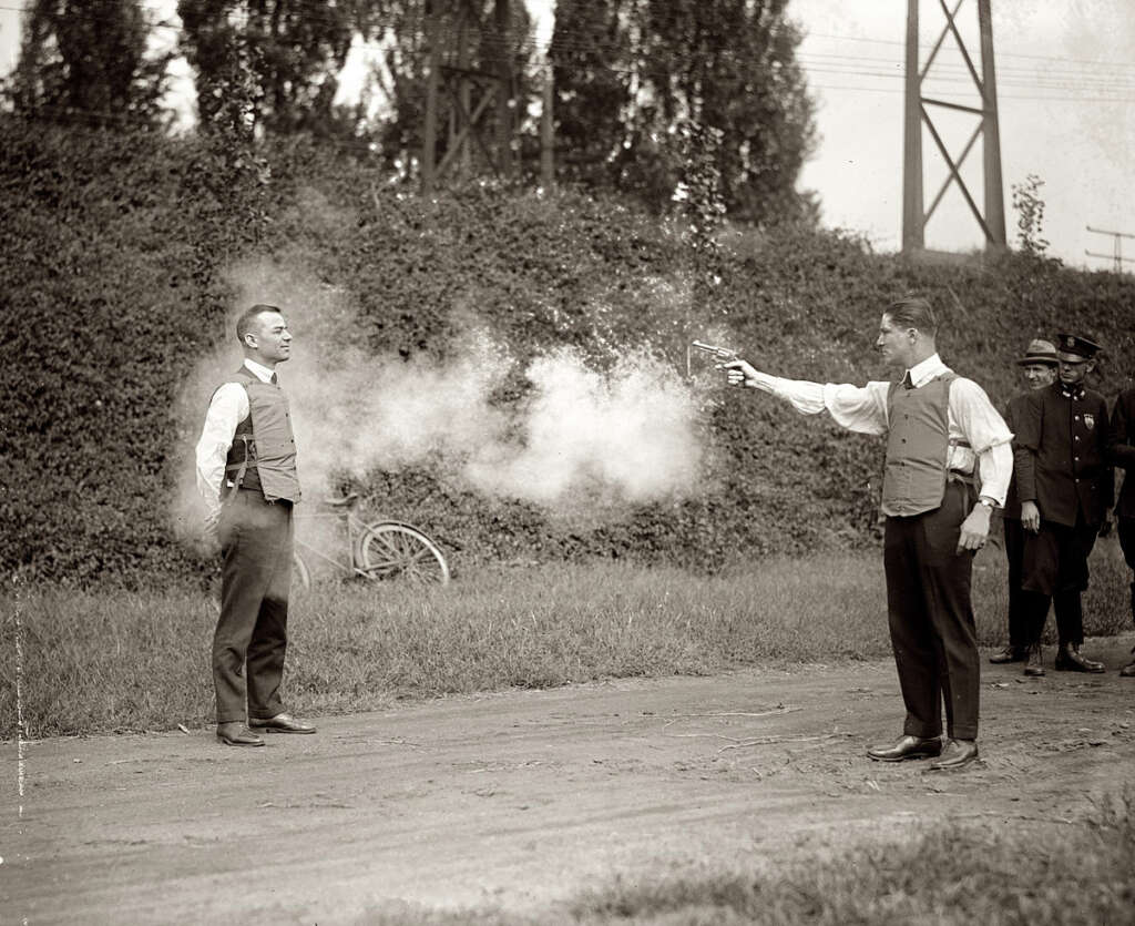 A vintage photo shows a man testing a bulletproof vest by firing a gun at another man wearing the vest. Smoke is visible from the gun discharge. Several observers, including policemen, watch from a distance. Trees and power lines are in the background.