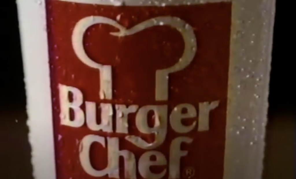 Close-up of a white cup with the Burger Chef logo, featuring a stylized chef's hat above the text. The cup has droplets of condensation on its surface. The background is dark and out of focus.