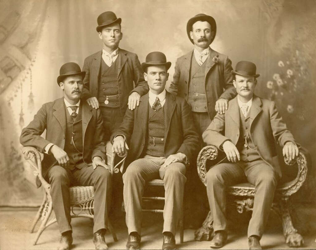 A sepia-toned vintage photograph of five men in suits and bowler hats. Three are seated in front, while two stand behind them. They pose formally in a studio with a painted backdrop.